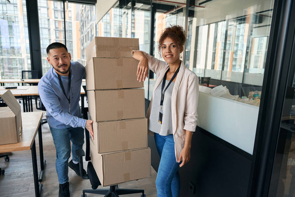 Pleased corporate worker and his happy female colleague posing for camera with pile of sealed cardboard boxes on swivel chair - Fotoğraf, Görsel