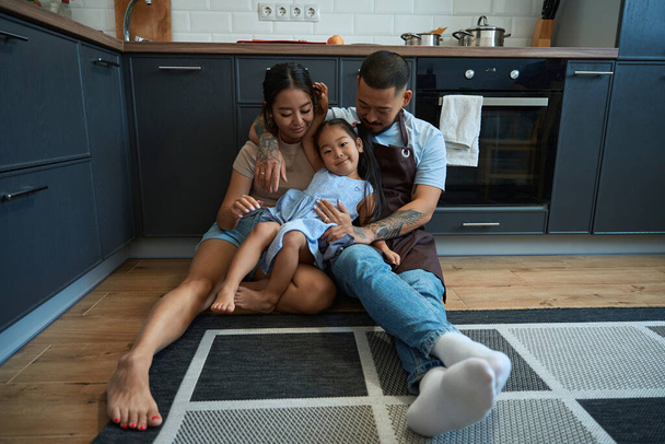 Ragazza crogiola tra le braccia dei suoi genitori, la famiglia si trova sul pavimento di una cucina accogliente - Foto, immagini