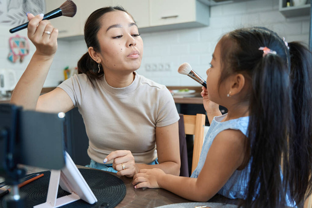 Mom blogger shoots a vlog, she and her pretty daughter hold makeup brushes in their hands - Photo, Image