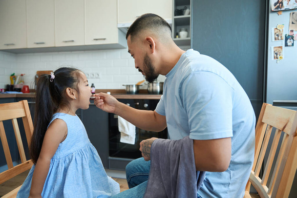 Mladý asijský otec dává poslušné dceři v modrých šatech lékařský sirup se speciální lžičkou - Fotografie, Obrázek