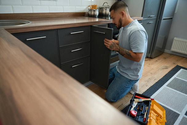 Young asian man in blue jeans crouched at the kitchen cabinet for minor repairs, next to the toolbox - Photo, Image