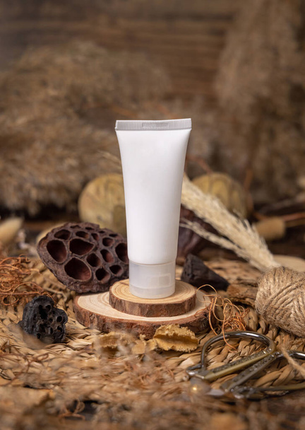 Biała plastikowa tuba kosmetyczna na drewnie w pobliżu naturalnych dekoracji zbliżenie, makieta opakowaniowa. Ekologiczny produkt do pielęgnacji skóry, balsam lub krem. Czeski skład z suszonymi liśćmi i kwiatami - Zdjęcie, obraz