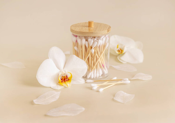 ライトベージュの白い蘭の花の近くの瓶の中に竹の綿の芽、閉じる。毎日の美容ルーチンのための有機スキンケア製品。環境に優しい生分解性綿棒 - 写真・画像