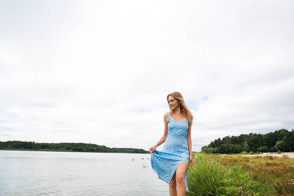 A beautiful slender girl with long blond hair, in a blue summer dress, walks on the water, in the river, against the backdrop of a picturesque landscape. - 写真・画像