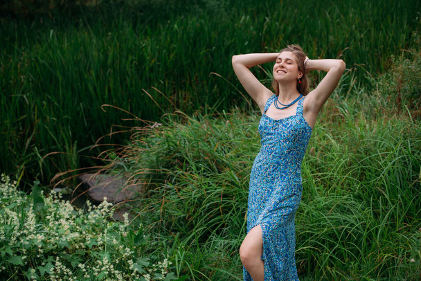 A slender young girl with blond hair, in a blue summer dress, stands on a green lawn in a forested area and smiles. - Foto, immagini