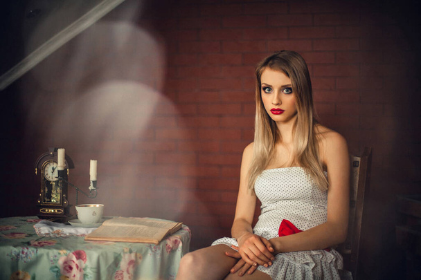 A gorgeous girl with bright make-up, with blond hair, in a light dress, sits in a room at a table. There is a candlestick with candles, a clock, a cup of coffee and a book on the table. - Фото, изображение