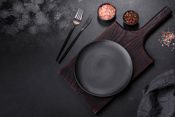 Пустая черная тарелка на темно-бетонном фоне со специями, травами и столовыми приборами. Домашний ужин - Фото, изображение