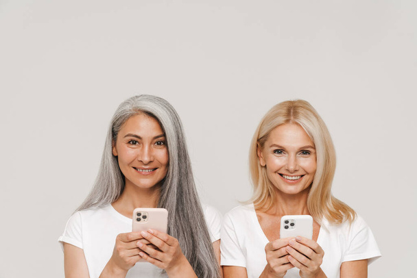 Mature multiracial women wearing t-shirts smiling and using cellphones together isolated over white background - Foto, Imagen