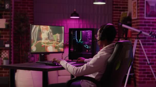 Statict tripod shot of african american gamer streaming first person shooter while explaining gameplay while using pc gaming setup. Man relaxing playing online action game talking to team on headset. - Footage, Video