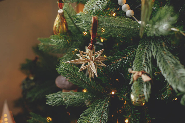 Christmas tree with vintage star and baubles with golden lights close up. Modern decorated christmas tree branches with stylish ornaments in festive room. Winter holidays, atmospheric time - Photo, Image