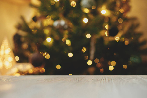 Tahta masa, Noel ağacının arka planında, altın ışıklı bokeh. Noel şablonu ve ürün için maket. Aydınlatılmış ağaçlı festival odasına karşı beyaz ahşap tahta - Fotoğraf, Görsel