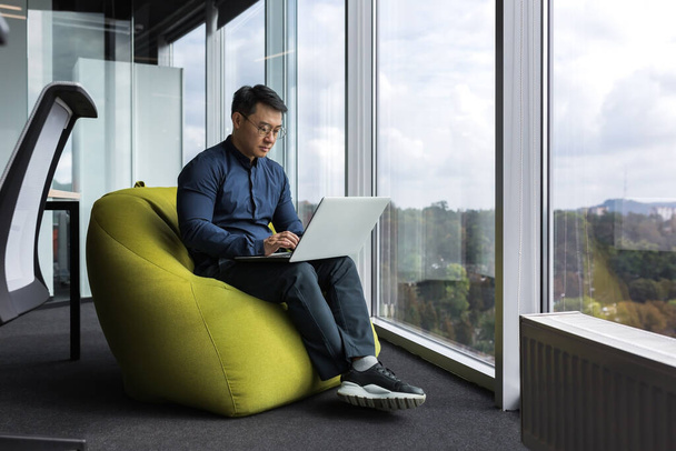 Asian young office worker sitting on soft ottoman chair, programmer using laptop for writing code and programming testing new program, man sitting near window inside office. - Foto, Bild