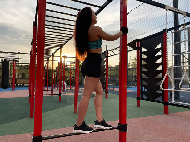 Full length shot of a long haired sportswoman resting on street workout playground after training on sunset - Foto, Bild