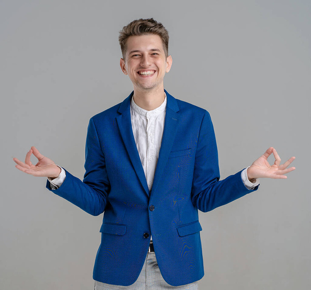 Portrait of a cheerful young man standing and showing ok gesture with two hands isolated over grey background. Waist up of young man in blue jacket - Fotografie, Obrázek