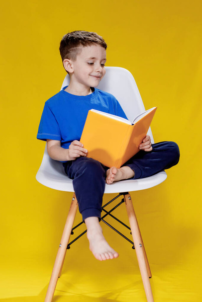 Little cheerful blond green-eyed boy 5-6 years old in a stylish blue T-shirt holding book and reading on yellow wall background, children's studio portrait. - Foto, afbeelding