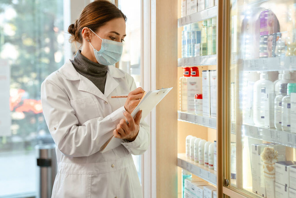 White apothecary wearing lab coat and face mask working in pharmacy indoors - Fotoğraf, Görsel