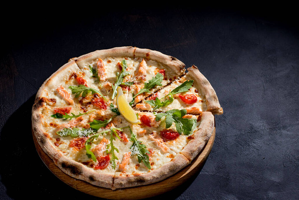 Pizza with salmon, mozzarella, cherry tomatoes, arugula, lemon and parmesan. Italian cuisine. On a black background. Free space for text. View from above - 写真・画像
