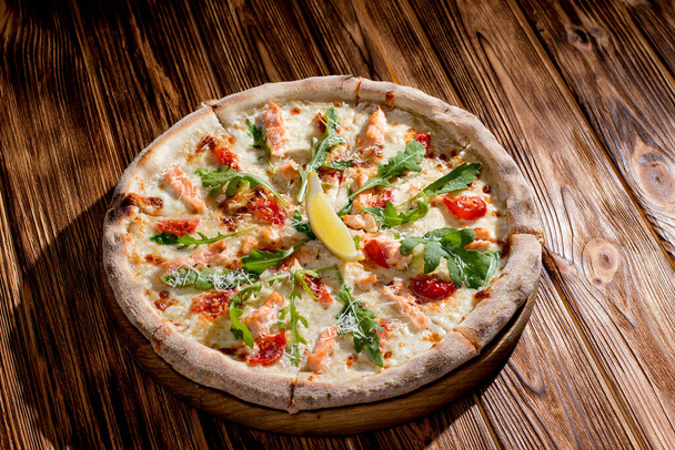 Pizza with salmon, mozzarella, cherry tomatoes, arugula, lemon and parmesan. Italian cuisine on a woden background. - 写真・画像