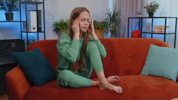 Displeased caucasian girl rubbing temples to cure headache problem, suffering from tension and migraine, stress, grimacing in pain, high blood pressure. Young child kid at home living room on couch - Footage, Video