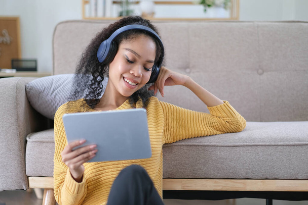 Photo of beautiful dark skin wavy lady homey mood holding digital tablet    earflaps listening new audio sitting floor near couch casual clothes yellow at living room indoor - Photo, Image