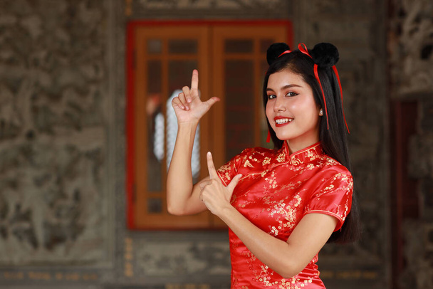 Beautiful Asian woman standing with red dress traditional cheongsam qipao and gesture of show something with happy smiling face on grey background (Chinese new year concept) - Photo, Image