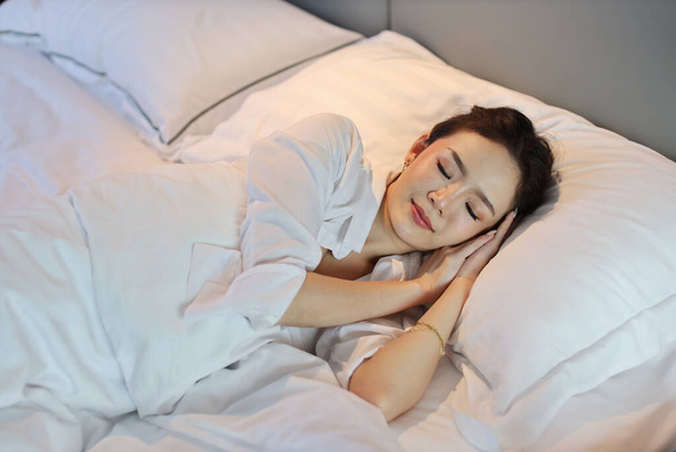 Beyaz uyku tulumu giyen Asyalı bir kadın yatakta yatarken ya da yatakta mutlu bir şekilde uyur ve güzel rüyalar görürken gülümserdi. Yaşam tarzı kavramı - Fotoğraf, Görsel