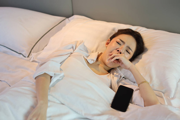夢を見る前に、白い寝衣を着たアジアの女性が寝そべって寝そべっていたり、携帯電話でベッドルームで寝ていたりするのを見渡します。ライフスタイルコンセプト - 写真・画像