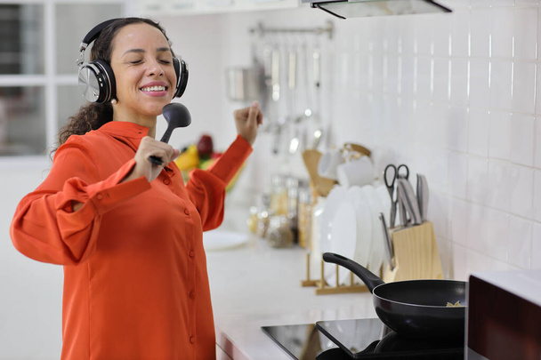 Feliz mujer latina cocinando, oliendo y saboreando comida mientras baila en la cocina. Hermosa joven hembra con auriculares preparando deliciosa comida mientras escucha música en casa. Concepto saludable y estilo de vida - Foto, Imagen