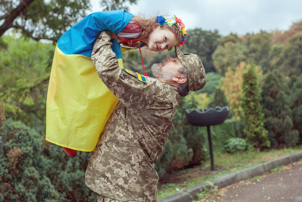 O exército ucraniano está segurando sua filha em seus braços, ela está envolvida na bandeira da Ucrânia. - Foto, Imagem