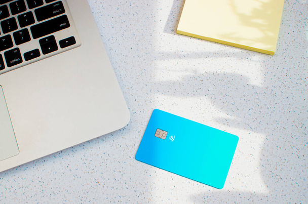 Creditcard met zichtbare chip en laptop, bovenop een tafel met zachte verlichting en schaduwen. Blauwe kaart op wit oppervlak. Concept: financiën, aankopen, betalingen, leningen, uitgaven, investeringen en schulden. - Foto, afbeelding