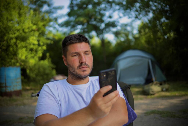 a man with a beard uses a phone in nature while waiting for a fish to bite the bait. in the background there is a fishing tent - Zdjęcie, obraz