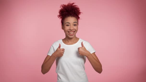 Muotokuva hymyilevä afroamerikkalainen punapää kihara nainen nostaa kädet näyttää kuin peukalot ylös näyttää kamera. Iloinen nainen valkoisessa t-paidassa eristetyllä vaaleanpunaisella taustalla kopiointitilalla. Ihmisten tunteita - Materiaali, video