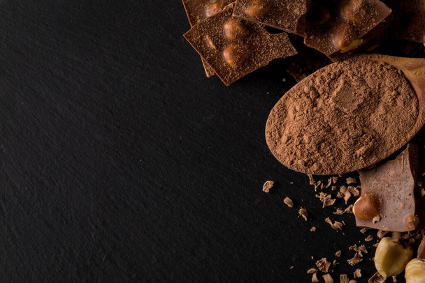 Разбитые кусочки шоколадных орехов и какао порошок в ложке на темном каменном фоне - Фото, изображение