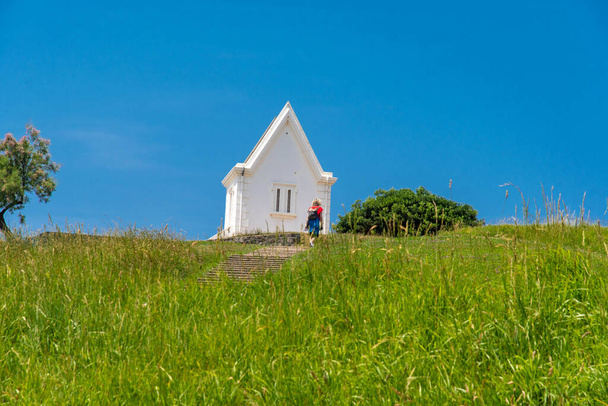 Malá bílá kaple na hoře se zelenou trávou a červeně oblečenou ženou - Fotografie, Obrázek