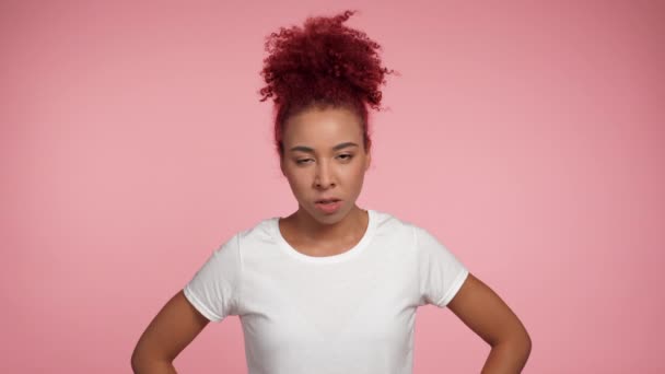 Közelíts meg egy dühös afro-amerikai vörös hajú göndör nőt, aki a kamerába néz. Portré gonosz nő visel fehér póló izolált rózsaszín háttér másolási hely. Az emberek érzelmek életmód koncepció - Felvétel, videó