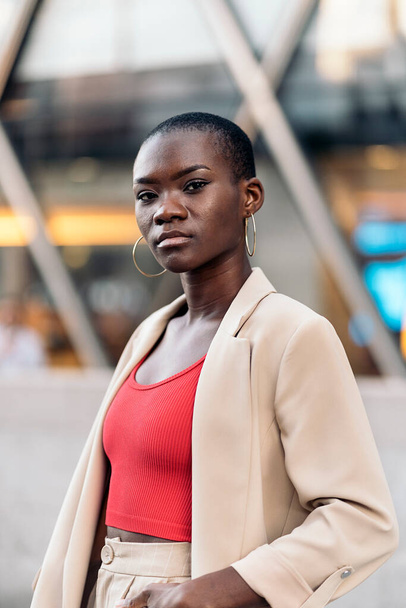 Vertical image of an elegant african american young adult woman with short hair standing and looking at camera in the city - Photo, Image