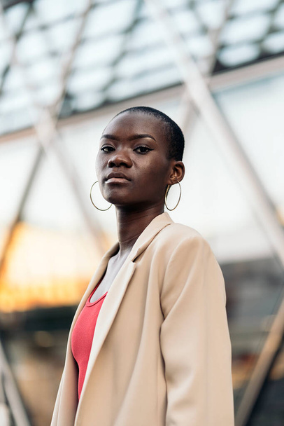 Low angle view of a stylish african american young adult woman with short hair standing and looking at camera in the city - Фото, изображение