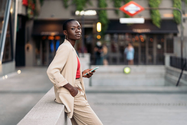 Side view of a stylish african american adult woman with short hair leaning on a concrete railing or small wall in the city with hand on pocket holding her phone while looking at camera - Foto, Imagem
