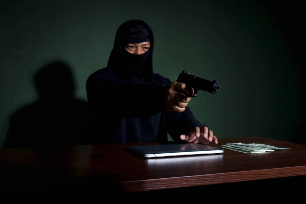 Homme criminel dans le capot noir tenant une arme à feu assis à un bureau avec ordinateur portable et de l'argent, voleur de masque commettant des crimes cybernétiques en ligne. - Photo, image