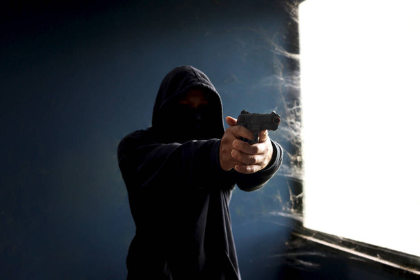 Εγκληματίας με μαύρη κουκούλα με όπλο να σημαδεύει το θύμα, κλέφτης μάσκας με το όπλο του στα χέρια για να διαπράξει έγκλημα σε εγκαταλελειμμένο κτίριο. - Φωτογραφία, εικόνα
