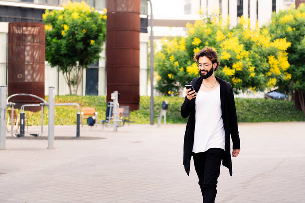 stylish young man with beard walking happy using a mobile phone in the city, concept of urban lifestyle and technology of communication, copy space for text - Photo, Image