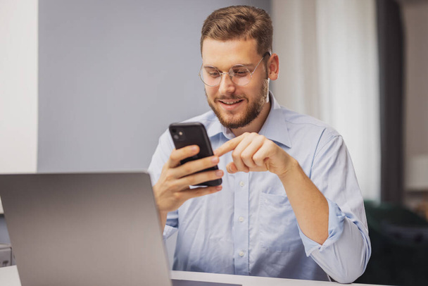 Молодий щасливий бородатий чоловік, використовуючи додаток для смартфона, сидить біля столу з ноутбуком вдома
 - Фото, зображення