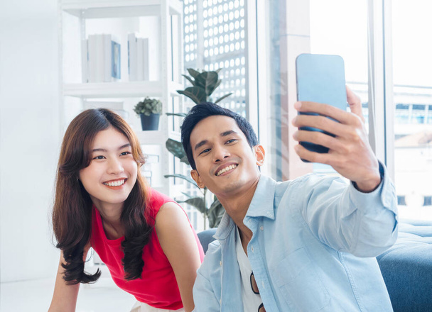 Happy handsome Asian man and young beautiful woman selfie while sitting near the glass window at white living room in high building. Cheerful couple taking portrait selfie with smart mobile phone. - Foto, Imagem