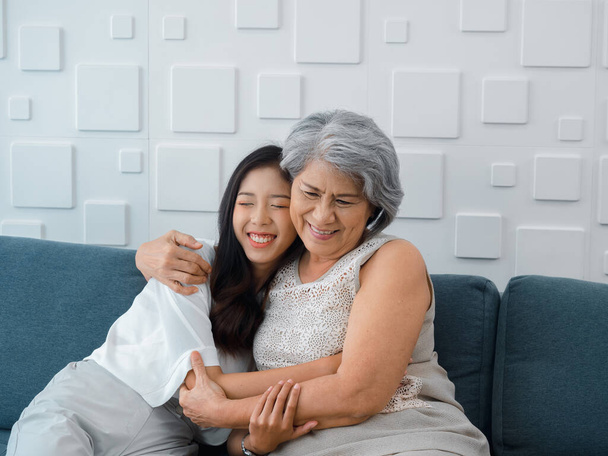 Porträt einer glücklichen asiatischen Seniorin, Mutter oder Großmutter mit weißem Haar, die ihre schöne Tochter oder ihr Enkelkind lächelnd umarmt und die Augen vor Liebe schließt, während sie zu Hause auf der grauen Couch im Wohnzimmer sitzt. - Foto, Bild