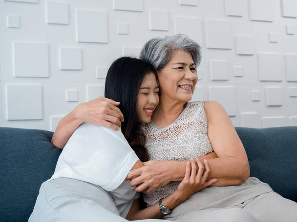 Portrét šťastné asijské senior, matka nebo prarodič bílé vlasy objímající svou krásnou dceru nebo vnouče s úsměvem a zavírající oči s pocitem lásky, zatímco sedí na šedém gauči v obývacím pokoji doma. - Fotografie, Obrázek