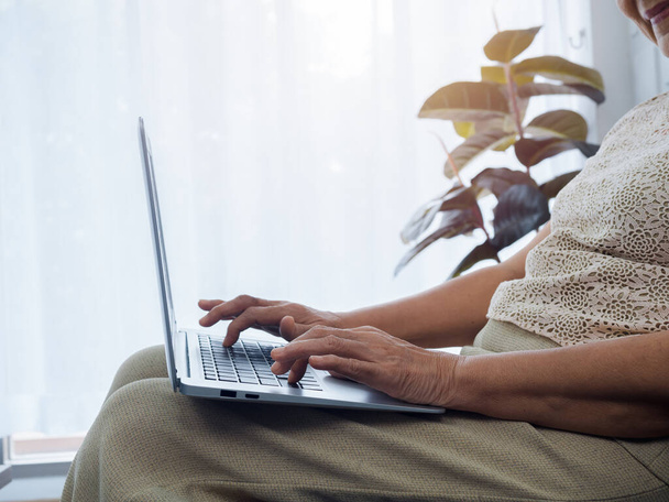 Κοντινά χέρια ηλικιωμένης γυναίκας που δουλεύει στο διαδίκτυο, χρησιμοποιεί φορητό υπολογιστή, κάθεται στον καναπέ στο σπίτι. Ηλικιωμένη γυναίκα ψάχνει κάτι στο διαδίκτυο. Ηλικιωμένοι με τεχνολογία. - Φωτογραφία, εικόνα