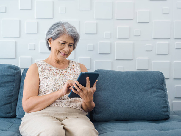 Mutlu Asyalı son sınıf öğrencisi kadın elinde dijital tablet ekranla oturma odasında oturuyor. Evde bilgisayar kullanan yaşlı bir kadın var. Yaşlılar teknolojiyi kolay kullanır.. - Fotoğraf, Görsel