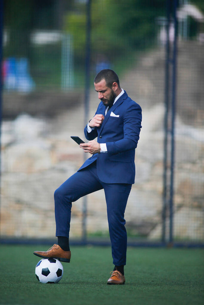 бізнесмен з м'ячем на нозі спілкується зі смартфоном всередині футбольного поля
 - Фото, зображення