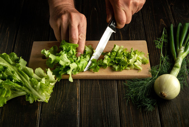 Las manos del chef cortan hojas verdes de lechuga en una tabla de cortar con un cuchillo para preparar comida vegetariana - Foto, imagen