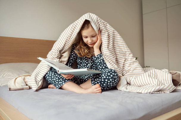 Cute little teen child girl lying and reading book on bed under the blanket  - Фото, зображення
